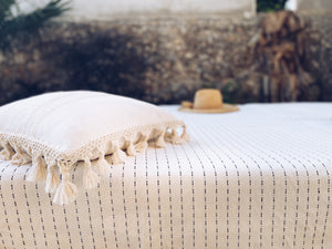 Natural cotton blanket with black dash embroidered pattern on a bed. Displayed with a pillow case with tassel and fringe.