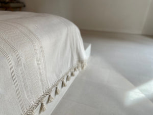 White natural colored handwoven bedcover displayed in a modern architectural room. Blanket has fringe and woven detail.