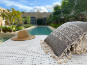 Collection of textiles poolside, featuring chambray pillow with fringe. Cotton handwoven pillow.