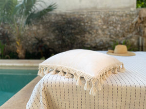 white pillow with fringe by pool with handwoven black and wide throw blanket and bedcover.